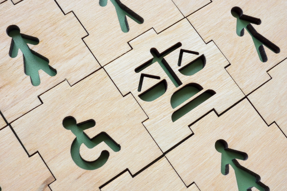 Wooden puzzle pieces with cutouts showing symbols for accessibility, people, and justice scales, representing disability rights and legal protection.