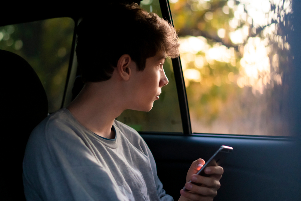 Teen sitting in a rideshare car at sunset on their phone, illustrating a rideshare child injury concern.
