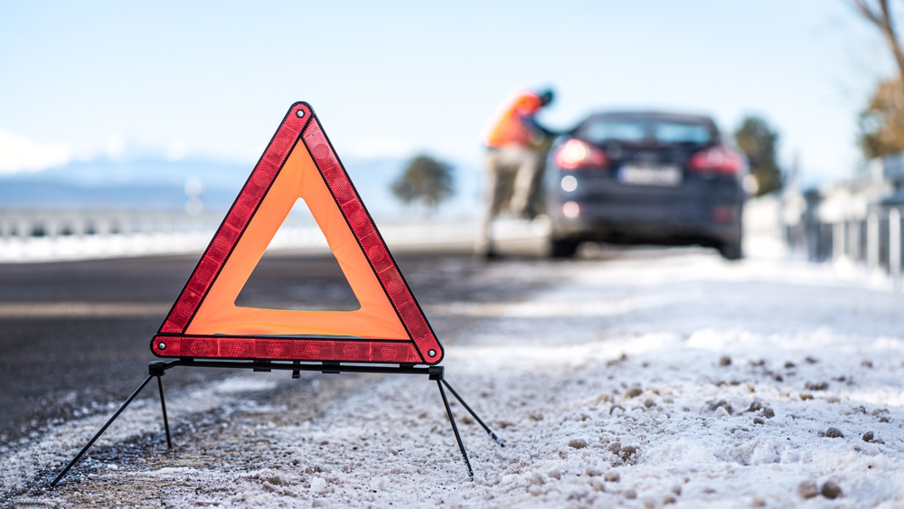 An orange emergency warning triangle on a snowy New Jersey roadside with a blurred car and driver in the background, highlighting the risks of holiday car accidents during winter.